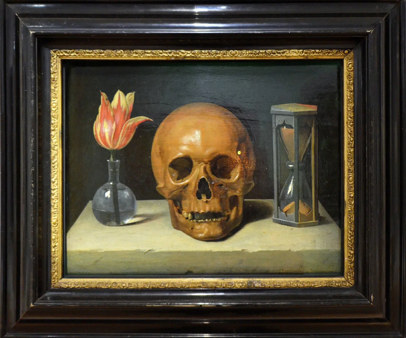 Tableau : un crâne posé sur une pierre, entre une tulipe dans un vase de verre et un sablier